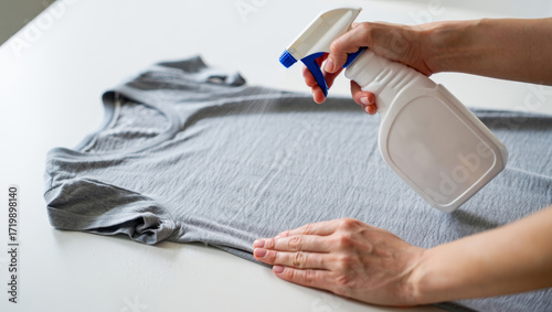 Hand using spray bottle on gray t shirt to remove wrinkles and freshen fabric, concept of quick clothing care, laundry solution, garment cleaning and home chores
