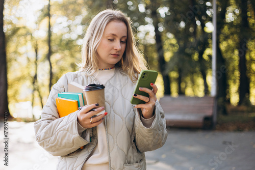 A young student woman walking in an autumn park, using her smartphone.