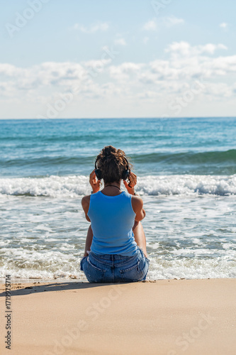 A woman is sitting on the beach with headphones on. She is listening to music and enjoying the ocean view