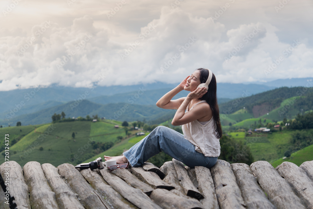 Naklejka premium A asian young woman sits on wooden logs in the mountains, wearing headphones, enjoying music and the scenic green valley view.