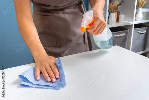 A person wearing a brown apron uses a spray bottle and a blue cloth to clean a white table surface in a home environment.