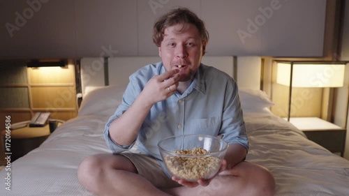 Cheerful traveler laughing out loud while watching a comedy film, hotel room, enjoying popcorn, traveler reacting with joyful amusement while watching tv movies