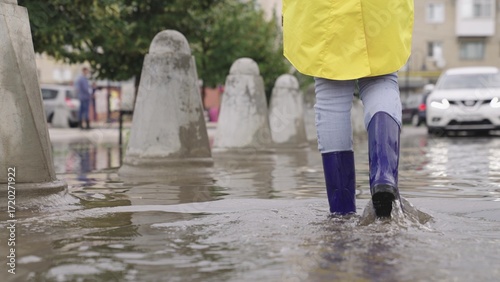Photos girls in rubber boots are walking in a puddle, a flood on a city street, flood on the sides of roads and asphalt roadway, splashes different directions from wet muddy puddle, presence water districts