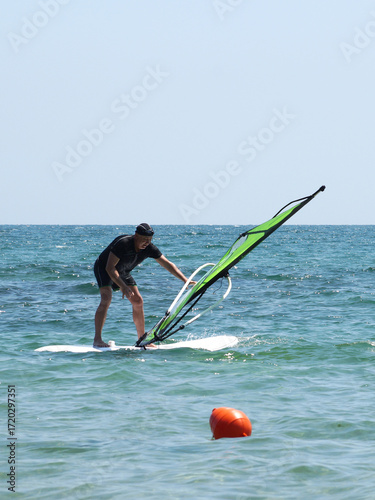 A man standing on a windsurfing board lifts a sail that fell into the water