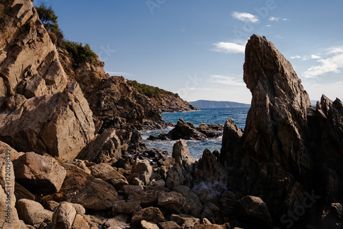 Dramatic seascape with jagged rocks rising from the clear blue water, waves crashing near a small sandy beach on the vibrant Greek coast.
