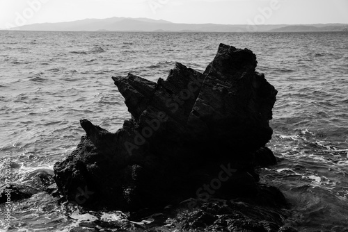 Dramatic seascape with jagged rocks rising from the clear blue water, waves crashing near a small sandy beach on the vibrant Greek coast.