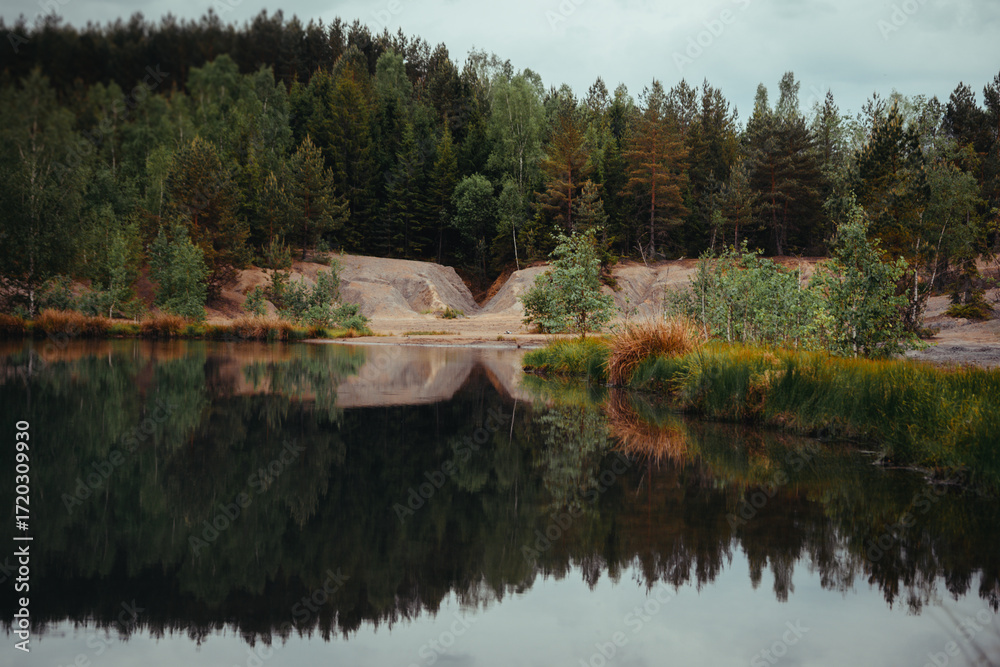 Obraz premium Serene Forest Lake Reflection. A tranquil lake reflects the lush green and brown trees of a surrounding forest under a slightly overcast sky.