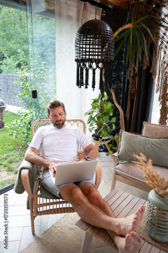 A bearded man sitting at home in a cozy interior, rubbing his tired eyes while working on a laptop. Exhausted Man Rubbing Eyes While Working on Laptop at Home