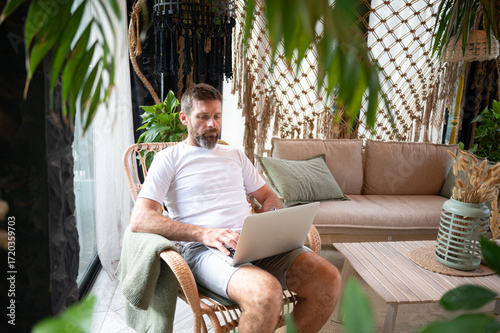 A bearded man sitting at home in a cozy interior, rubbing his tired eyes while working on a laptop. Exhausted Man Rubbing Eyes While Working on Laptop at Home