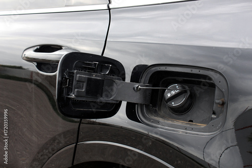 Close-up of a black car fuel filler area with open gas cap cover. The matte black cap contrasts with the glossy painted body. Diesel Tank Hatch, Gasoline Tank Hatch, Fuel Tank Hatch is open in car.
