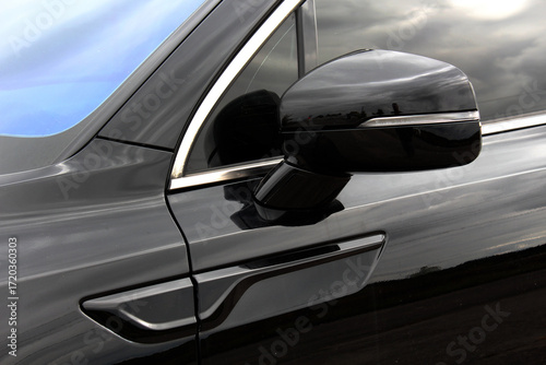 Rearview mirror on a black car. Close-up of glossy black car door, featuring a side mirror with chrome trim. Road safety while driving. Black car mirror. Side rear-view mirror lux car.
