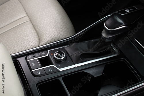 Black car center console with leather gear shifter, rotary drive mode selector, and control buttons, accented by silver trim and beige patterned seat upholstery.