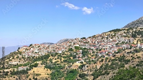 Fototapeta Naklejka Na Ścianę i Meble -  Panoramic view of Arahova, Greece, a picturesque mountain town on the slopes of Mount Parnassus, with red-tiled houses, stone buildings, and lush greenery under a clear blue sky.