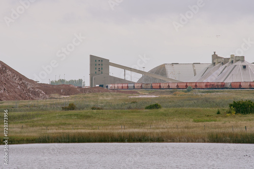 Potash factory with covered mineral piles and rail cars near a water body under cloudy skies in Saskatchewan, Canada.