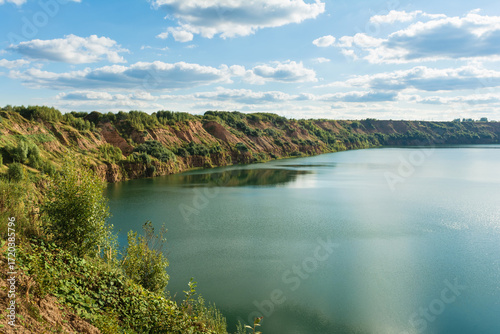 A calm lake surrounded by green, rugged cliffs under a bright blue sky with scattered clouds, showcasing a peaceful and scenic natural landscape.