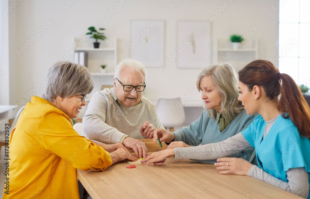 Obraz premium Happy smiling senior elderly people group sitting at table in retirement home collecting geometric figures with young friendly nurse. Dementia prevention and leisure time in nursing home concept.