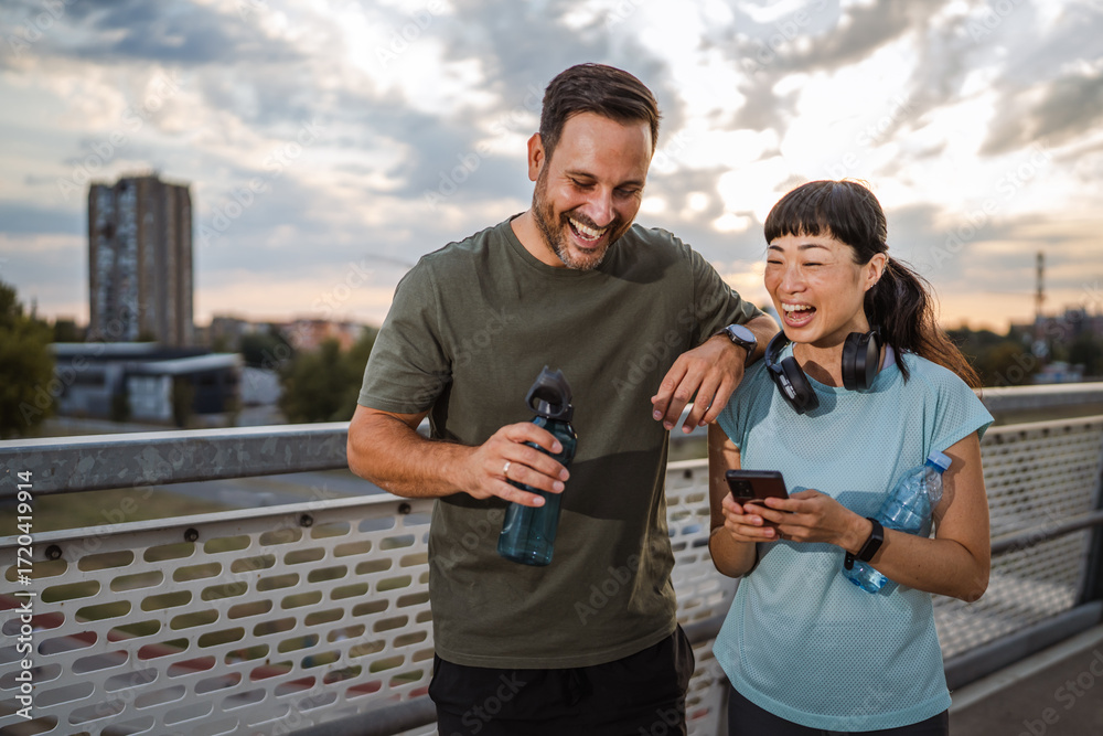 Fototapeta premium Diverse couple laughing and checking smart phone after urban exercise