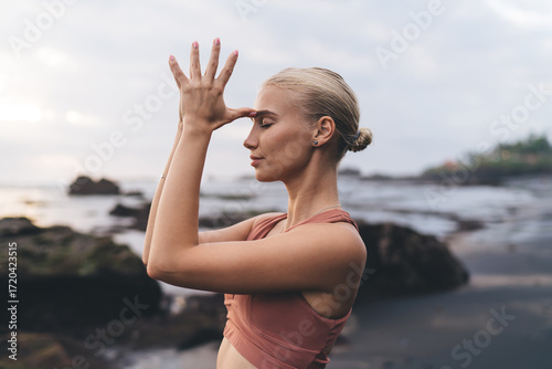 Photos Soft close-up of woman practicing yoga mudra with fingers to third eye, eyes shut, standing against ocean at dusk, symbolizing personal connection, spiritual vision and post-digital awareness