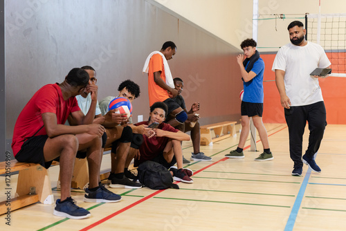Diverse male volleyball team on wooden bench holding volleyballs in gym while coach walking with tab