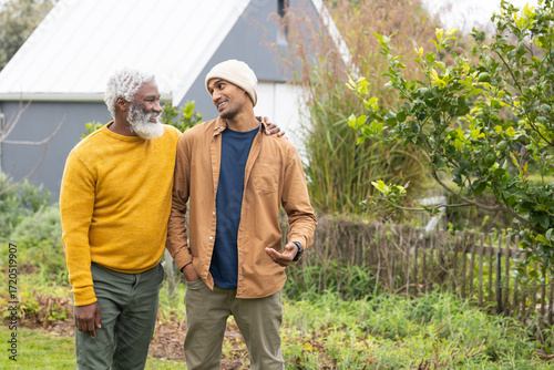 Diverse father and son standing in garden beside gray-sided outbuilding with wooden picket fence