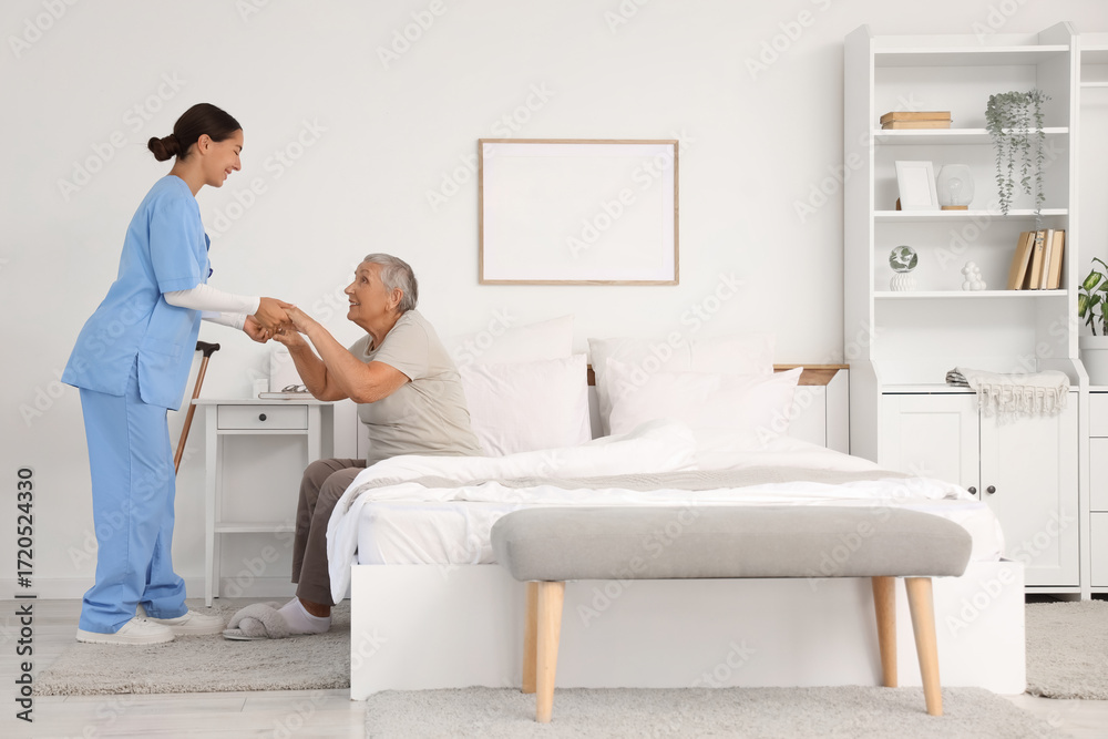 Fototapeta premium Female caregiver helping happy senior woman to stand up in bedroom
