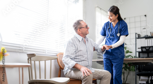 A friendly female nurse in blue scrubs stands beside an elderly male patient, offering him comfort and support. The clinic or home setting suggests a moment of compassionate healthcare,