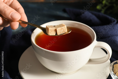 Tableau sur toile Young woman adding lumps of sugar into cup of black tea on blue napkin as backgr