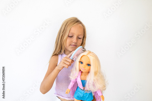Little blonde girl with carefully combs the hair of a blonde fashion doll. She is focused and engaged in creative play on a white background. Traditional children's hairdresser game for girls.