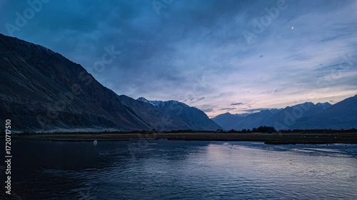 timelapse of Himalayas and blue sky mirrored in the Shyok River at sunrise, near lush Sumur village—where green orchards and barley fields thrive against Nubra Valleys stark mountain beauty.