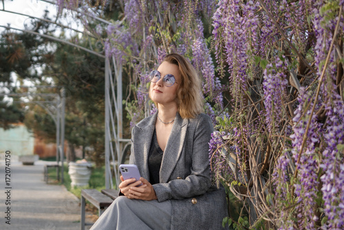 Woman Wisteria Garden Bench - A woman sits on a bench in a garden with blooming wisteria vines.