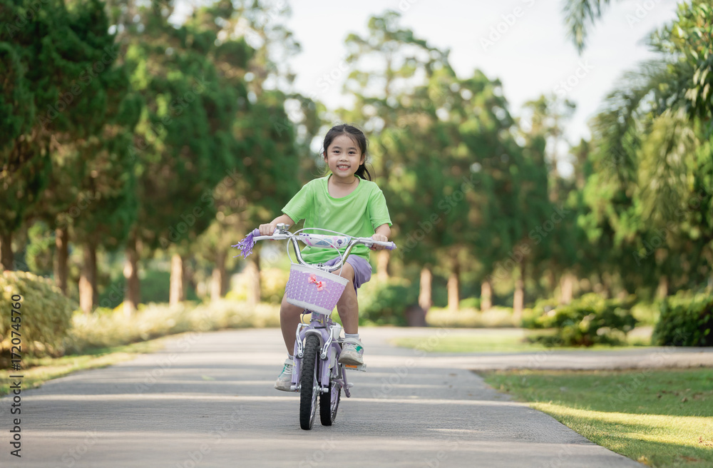Fototapeta premium Happy young girl riding bicycle in park during sunny day, enjoying childhood and outdoor activities in nature, healthy lifestyle and joy of movement