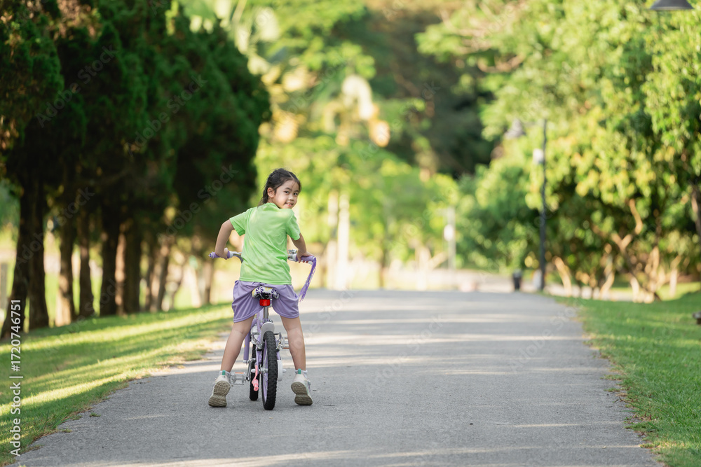 Obraz premium Young girl riding bicycle in park, enjoying sunny day, smiling with joy, greenery surrounding, outdoor activity, childhood fun, carefree moments
