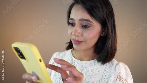 Closeup of happy smiling beautiful young Indian girl woman hold phone using mobile typing sms online chatting feels happiness, isolated over grey background.