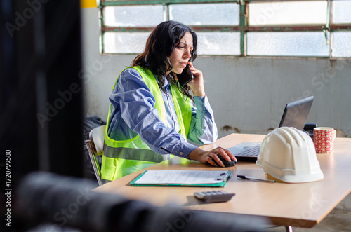 Young female employee using a mobile phone and laptop working at warehouse industry office. Deposit factory concept.