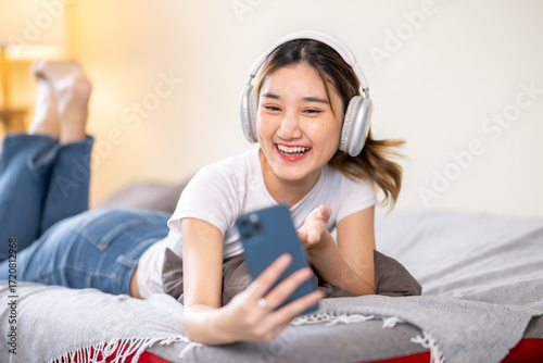 Happy asian woman wear headphones video calling talking on phone while lying on sofa bed in a room.