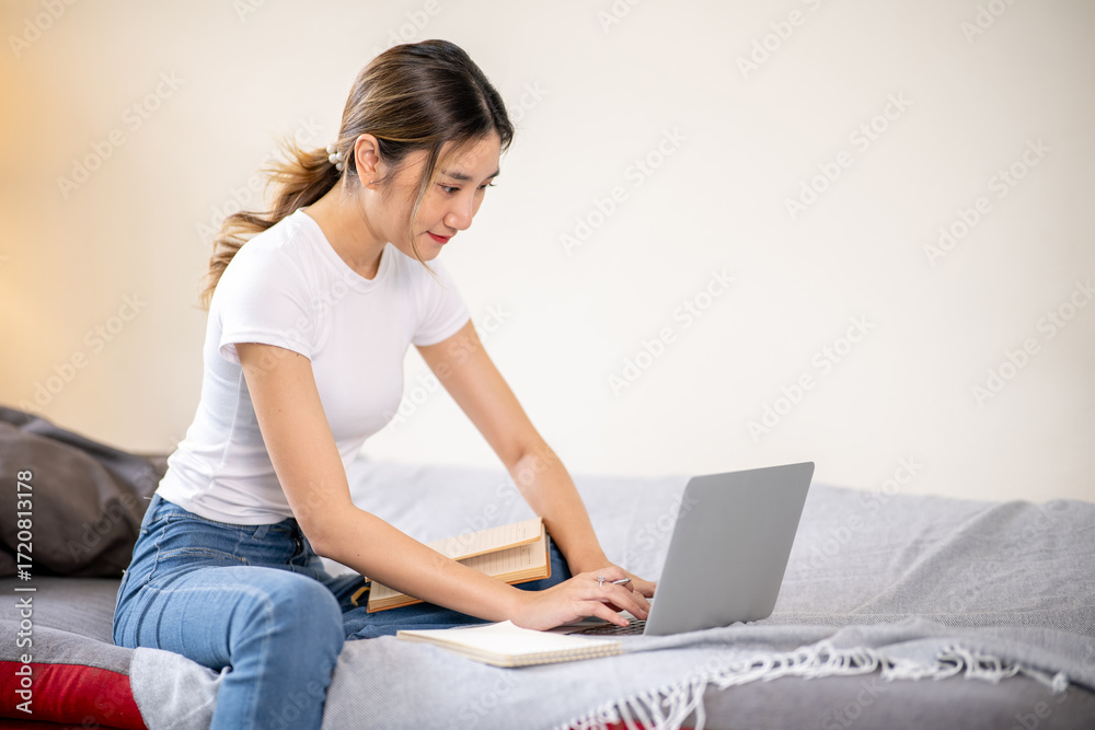 Naklejka premium Asian woman looking and typing on laptop aside books while sitting on sofa bed in a living room.