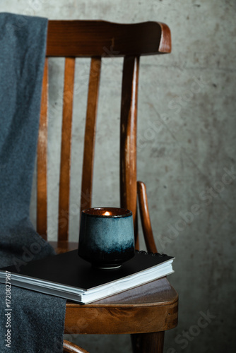 Lit candle in ceramic cup on notebook with wooden chair and gray fabric. Cozy minimal still life in wabi-sabi mood.
