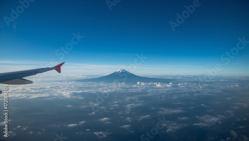 A high-altitude view of a majestic mountain peak, Fuji, from an airplane window, with a vibrant blue sky and a vast expanse of clouds below.