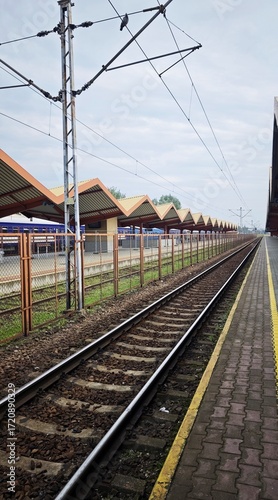 Empty railway tracks at a train station with patterned roofs and electric lines under a cloudy sky