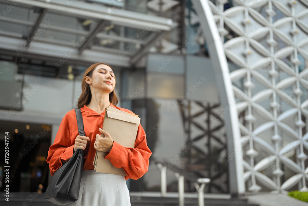 Fototapeta premium Asian businesswoman smiling peacefully, holding documents while standing outside a modern office building