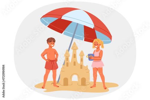 Children build sand castle on beach, standing with umbrella to protect them from sun. Little positive boy and girl play together on beach during summer trip to sea or ocean with parents