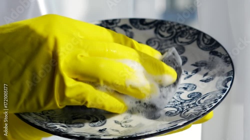 Protecting your hands while washing dishes.
Cleaning up the kitchen after dinner and washing dishes.
A housekeeper wearing yellow gloves washes a dirty plate in a kitchen with daylight.