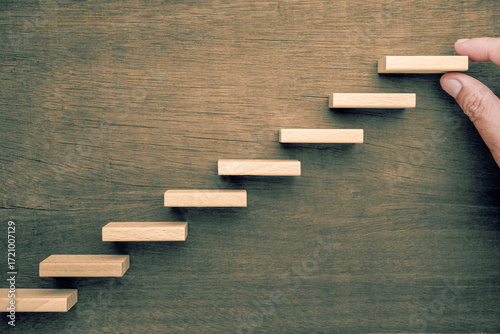Wooden blocks arranged as upward steps on wooden background with hand placing the top block representing continuous improvement and personal development in lifelong learning
