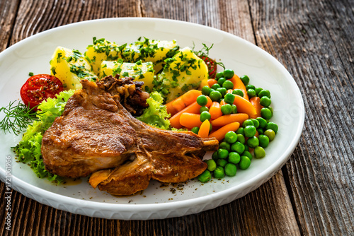 Baked veal cutlet with bone, boiled potatoes, mini carrots and green peas on wooden table