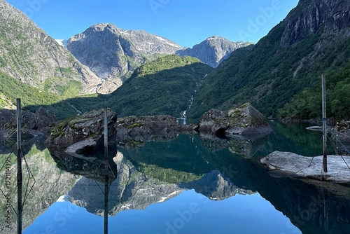 Panoramic landscape view of a mirrored surface of Bondhusvatnet lake, reflecting mountains and a waterfall, near Odda in Norway	