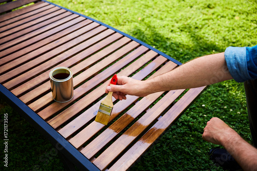 Man oiling wooden sunbed with paintbrush in backyard garden