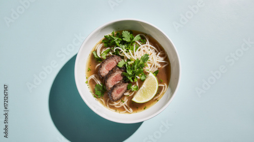 Pho noodle soup with rare beef in a white bowl on a pastel aqua background, clean slight overhead studio shot