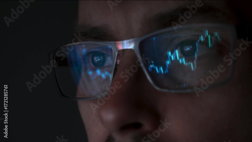 Close up view of focused trader wears eyeglasses looking at pc screen with stock market charts reflection, working online late night. Man analysing crypto market. Selective focus on eye