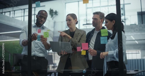 Employees Engage in a Creative Planning Session, Writing On and Placing Post-It notes on a Glass Wall to Structure a Business Project. They Discuss Each Suggestion and Rearrange Notes