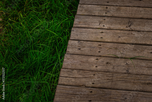 A close-up view of a weathered wooden boardwalk meets vibrant green grass, highlighting the contrast between natural and built paths.